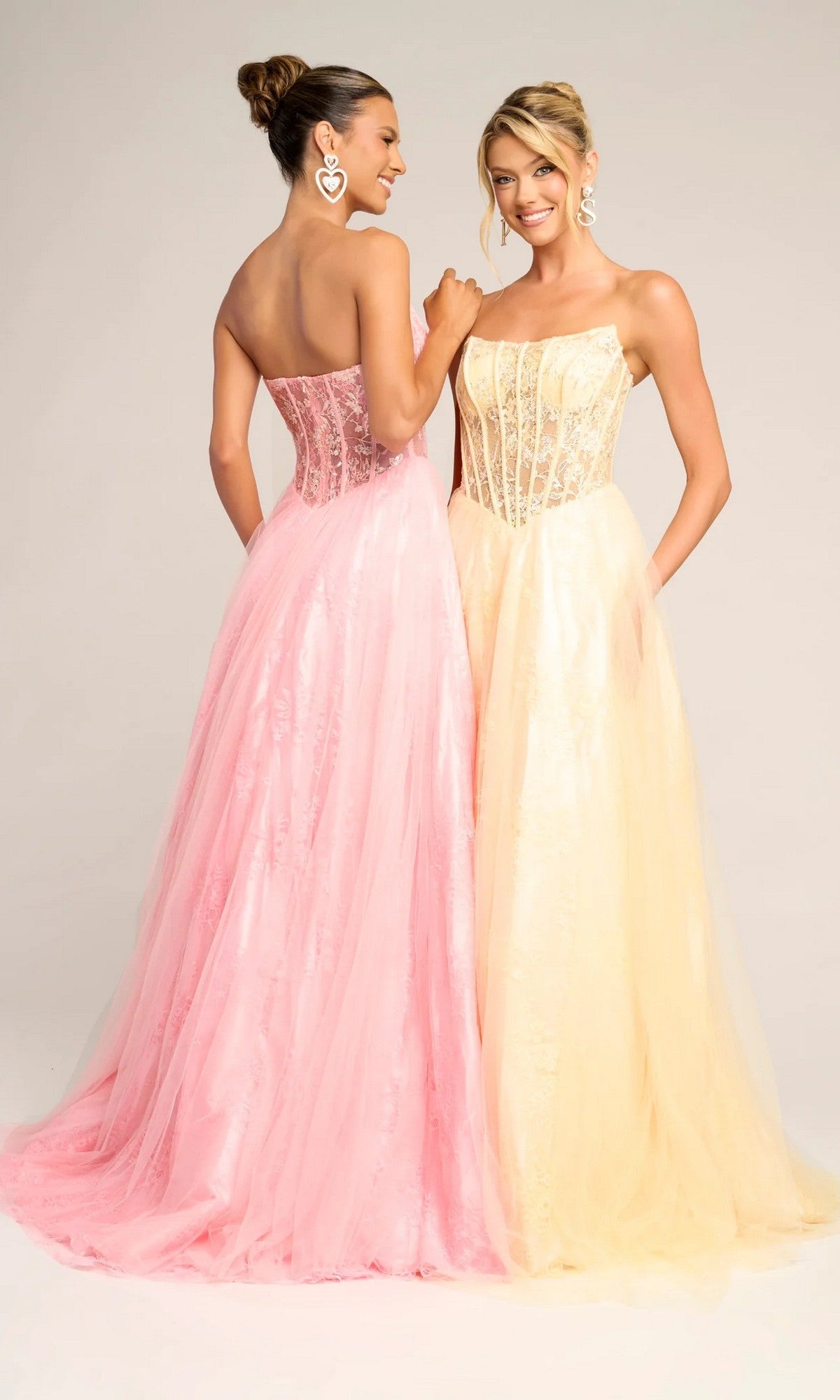 Two women stand side by side against a plain background, smiling in Portia and Scarlett XO023 long formal dresses—one wears pink. Each strapless gown features a lace corset bodice with tulle and elegant lace details.